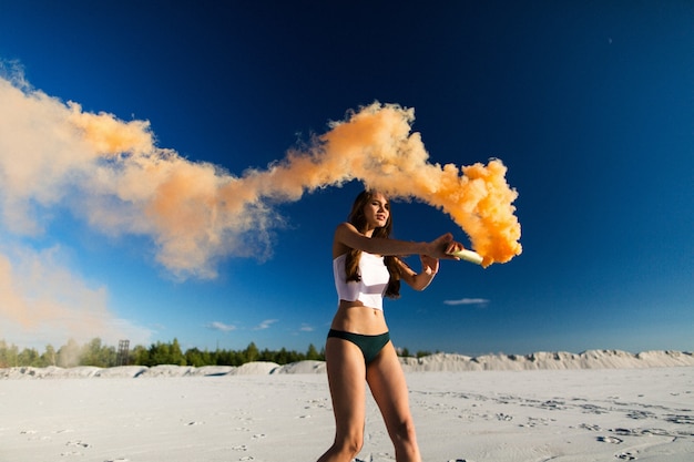 Woman walks with orange smoke on white beach under blue sky