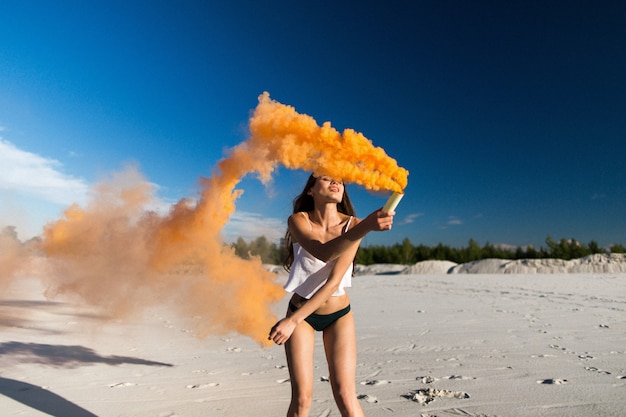 Foto gratuita la donna cammina con il fumo arancione sulla spiaggia bianca sotto il cielo blu