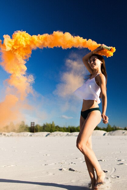 Woman walks with orange smoke on white beach under blue sky