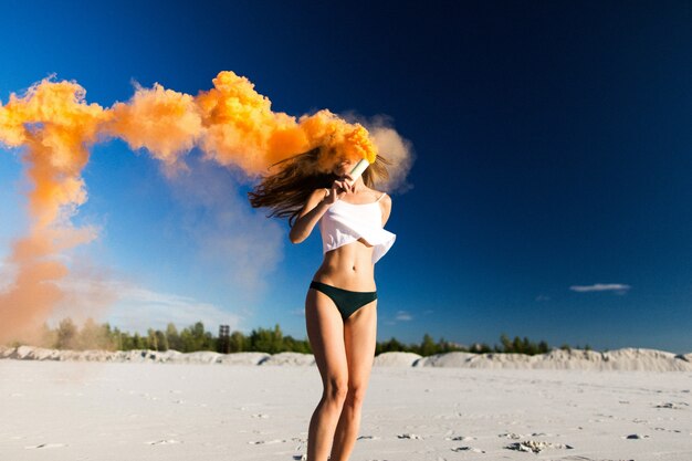 Woman walks with orange smoke on white beach under blue sky