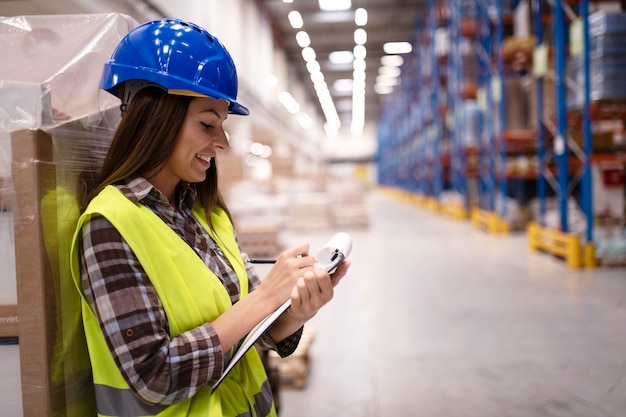 Woman warehouse worker leaning on cardboard boxes and taking notes in large warehouse distribution center