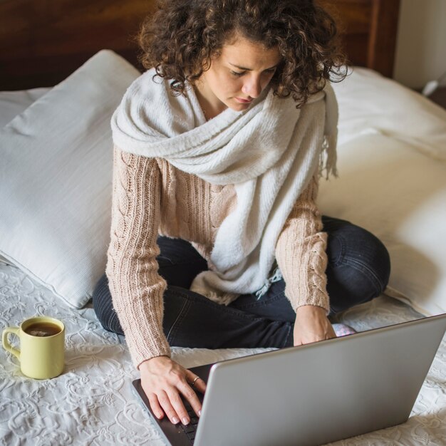 Woman in warm clothes using laptop on bed