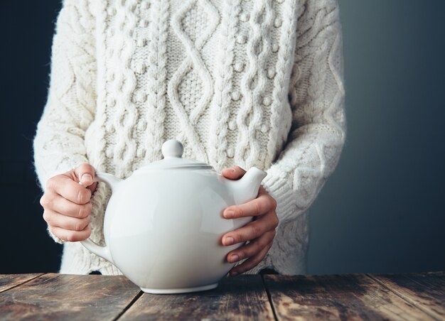 Woman in warm knitted thick sweater holds big white teapot with tea on grunge wooden table. Front view, anfas, no face.