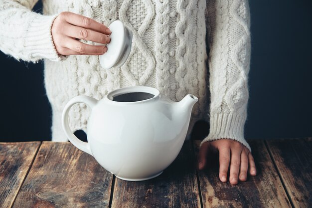 Woman in warm knitted thick sweater holds cap of big white teapot in hands on grunge wooden table. Front view, anfas, no face.