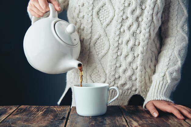 Woman in warm knitted thick sweater pours black tea from big white teapot to cup on grunge wooden table. Front view, anfas, no face.