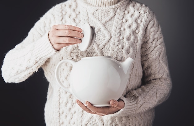 Woman in warm sweater holds big white ceramic teapot on one hand and opens cap with other
