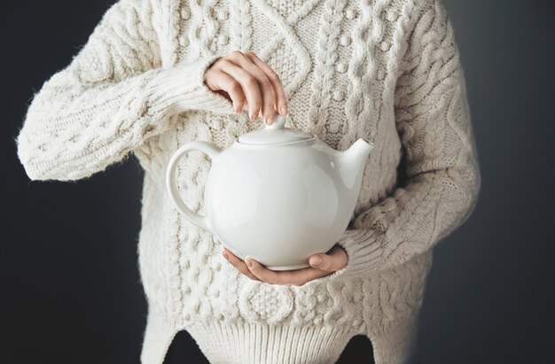 Женщина в теплом свитере держит большой белый чайник с чаем в форме сердца. Вид спереди, деревянный стол гранж. анфас, без лица.
