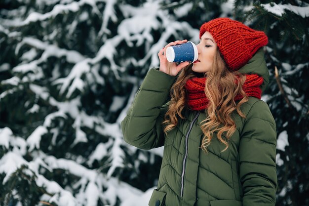 暖かい冬の服の女性は、冬の雪の日に屋外でコーヒーを飲みます。