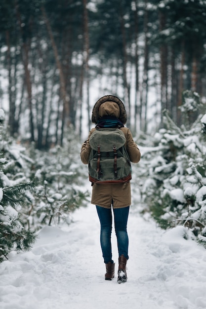 Woman in warm winter jacket with fur hood and big travel rucksack in snowy winter forest