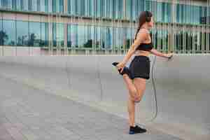 Free photo woman warms up before jogging raises leg stretches muscles dressed in sportswear prepares for cardio training focused forward poses near modern city glass building