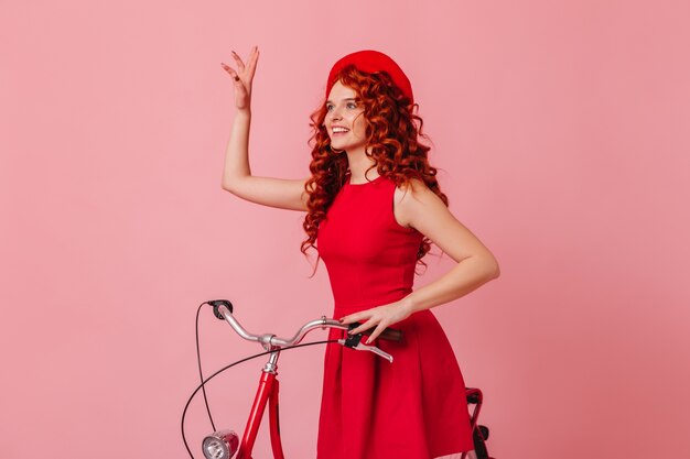 Woman waving greeting while sitting on bicycle on pink space. lady in red beret and dress.