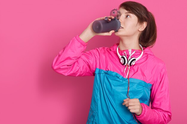 Woman wearin blue and rose sportswear drinking water from bottle, model posing isolated on rosy. young female fitness instructor or personal trainer in studio.