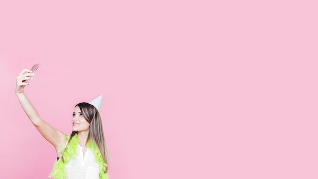 Free photo woman wearing boa and party hat taking selfie against pink background