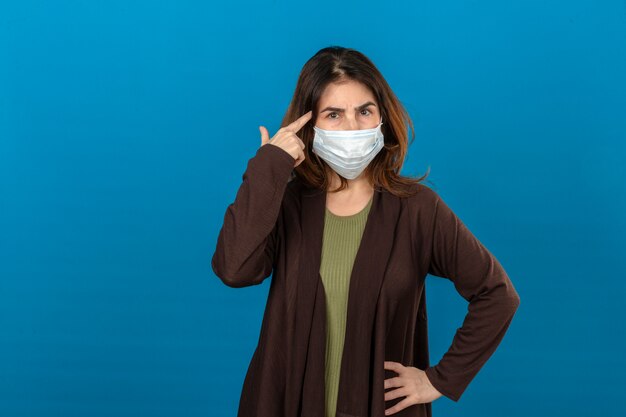 Женщина в коричневом кардигане в медицинской защитной маске, указывающая на храм с мышлением пальцем, сосредоточилась на задаче с хмурым лицом над изолированной синей стеной
