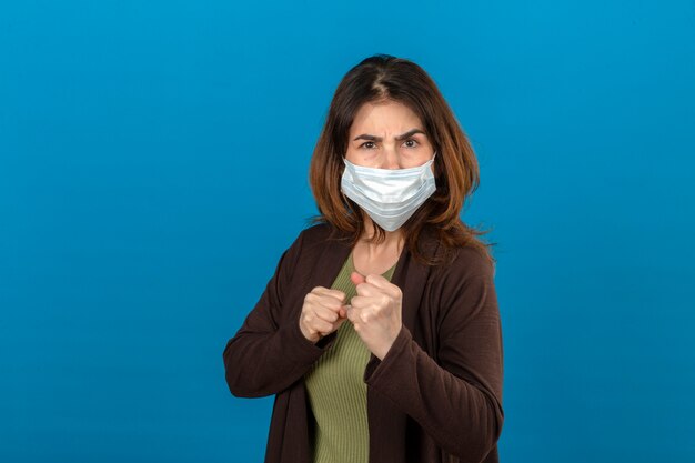 Woman wearing brown cardigan in medical protective mask standing with boxing fists looking and ready to attack or defense