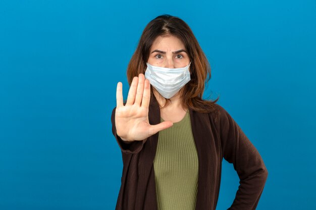 Женщина в коричневом кардигане в медицинской защитной маске стоит с открытой рукой и делает знак остановки с серьезным и уверенным выражением лица, защищая жест над изолированной синей стеной