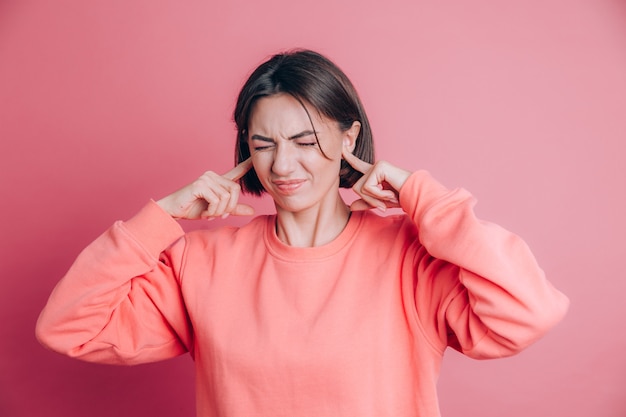 Женщина в повседневном свитере на фоне закрывает уши пальцами с раздраженным выражением лица из-за шума громкой музыки.