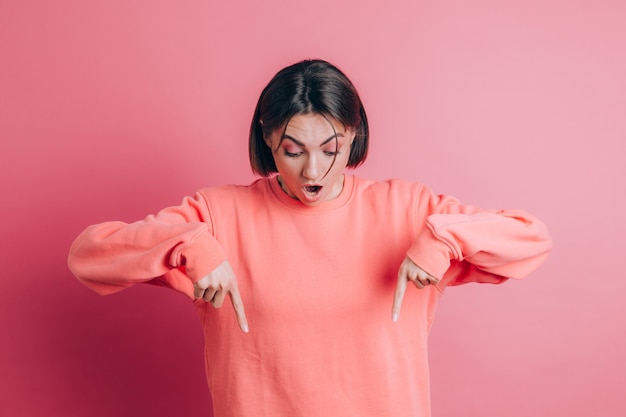 광고, 놀란 얼굴과 오픈 입을 보여주는 손가락으로 아래를 가리키는 배경에 캐주얼 스웨터를 입고 여자