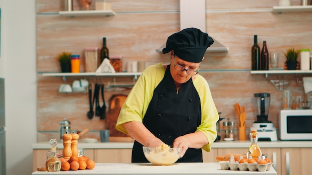 Женщина в костюме шеф-повара, смешивая треснувшие яйца с мукой на кухне, готовит еду по традиционному рецепту. Пожилой пекарь на пенсии замешивает в стеклянной миске ингредиенты для домашнего торта