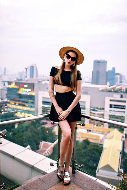 Woman wearing crop top and mini skirt, straw hat and trendy accessories, posing at roof top of luxury hotel in big modern city