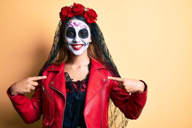 Woman wearing day of the dead costume over yellow looking confident with smile on face pointing oneself with fingers proud and happy