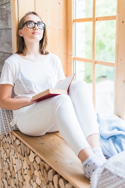 Foto gratuita occhiali d'uso della donna con il libro che si siede vicino alla finestra