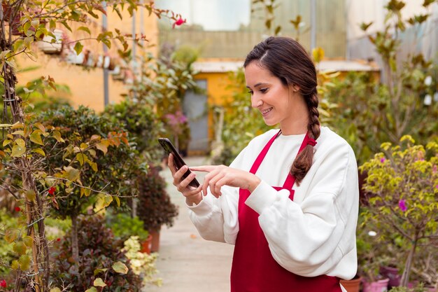 Woman wearing gardening clothes and holding phone in greenhouse