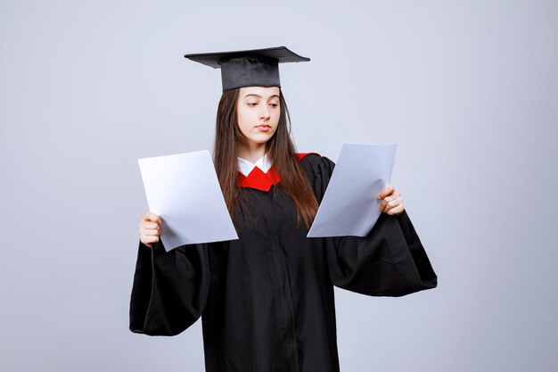 Woman wearing graduation cap and ceremony robe holding empty papers. High quality photo