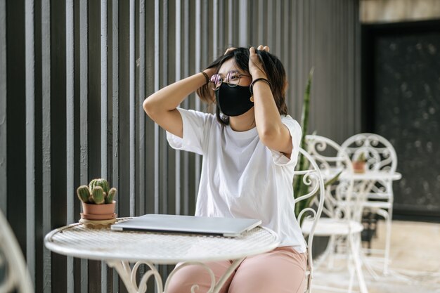 Женщина в маске играет на ноутбуке и обеими руками держит голову