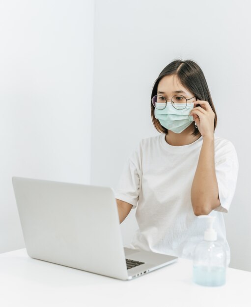 A woman wearing a mask playing a laptop and having a bottle of handwashing gel.