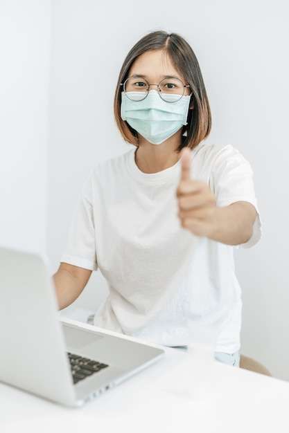 A woman wearing a mask playing a laptop and thumbs up.