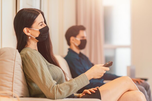 남자 친구와 함께 호텔 로비에 앉아 있는 동안 원격 제어를 사용하여 의료 마스크를 쓴 여자. 관계, 여행, 코로나바이러스, covid19 개념