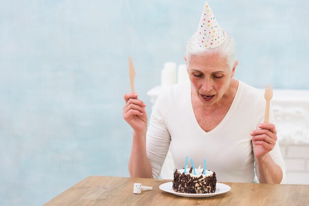 Woman wearing party hat holding wooden knife and fork looking at birthday cake on table