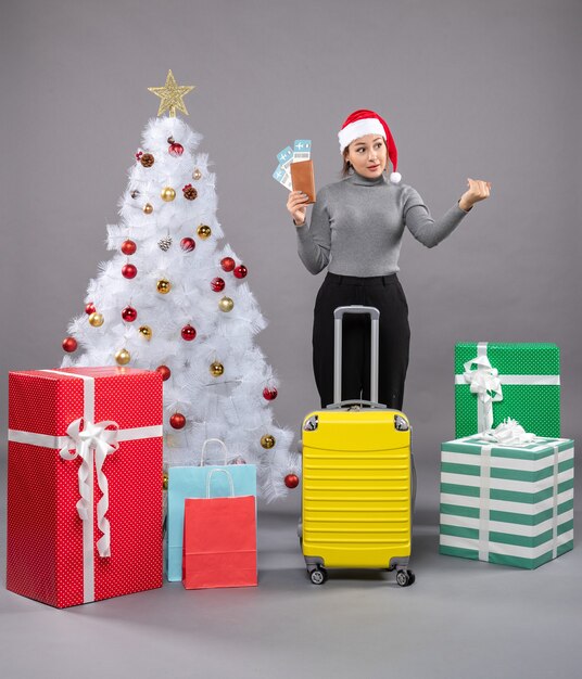 Woman wearing Santa Claus hat with luggage next to Christmas tree