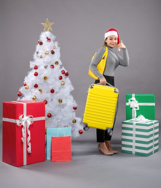 Woman wearing Santa Claus hat with luggage next to Christmas tree