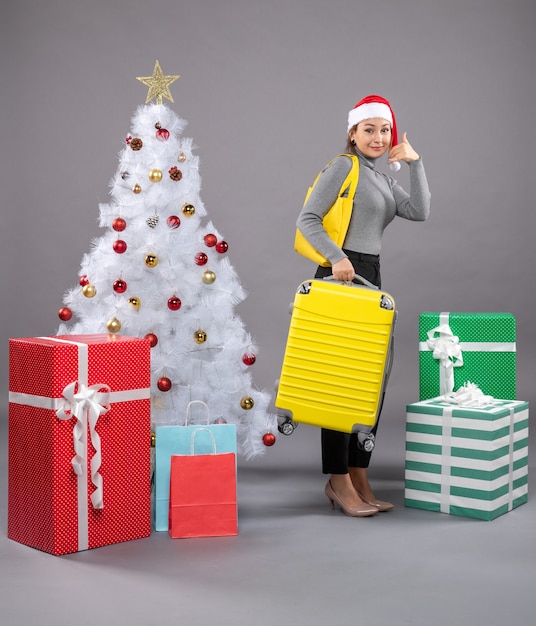 Woman wearing Santa Claus hat with luggage next to Christmas tree