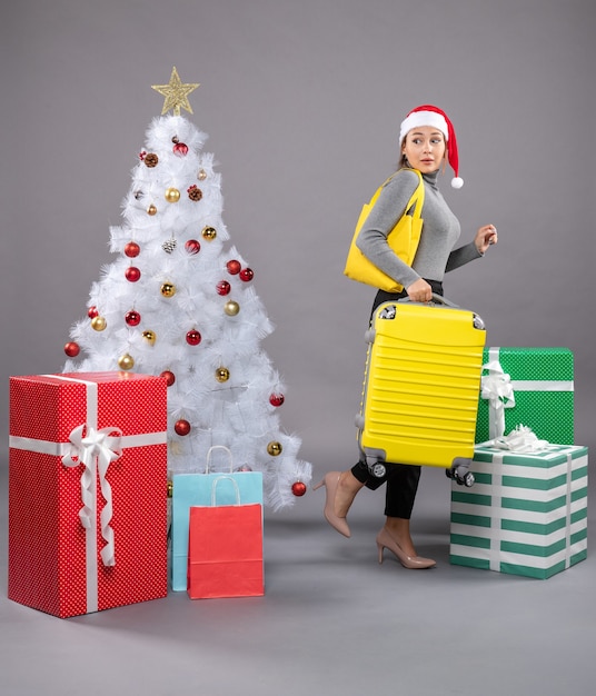 Woman wearing Santa Claus hat with luggage next to Christmas tree