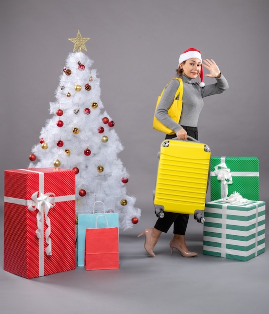 Woman wearing Santa Claus hat with luggage next to Christmas tree