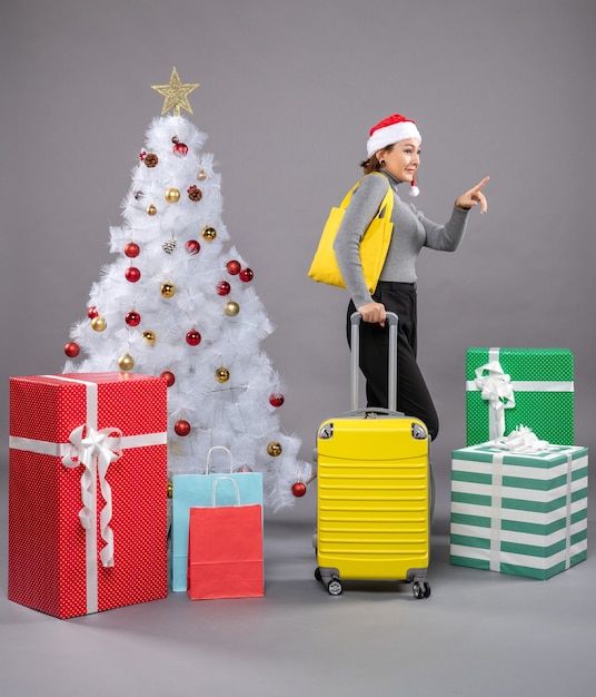 Woman wearing Santa Claus hat with luggage next to Christmas tree