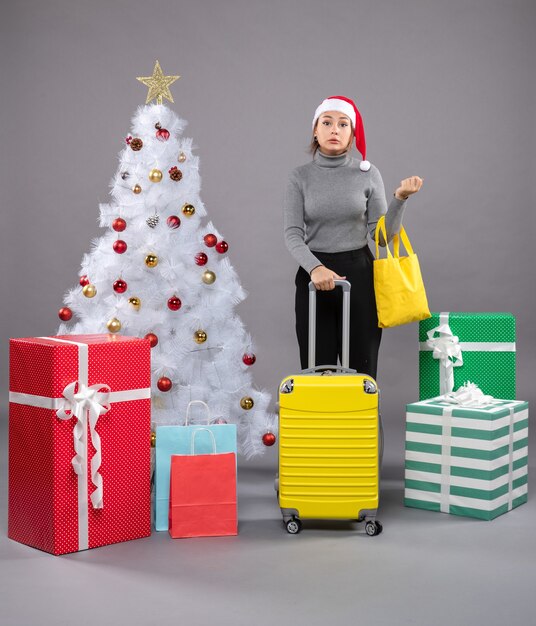 Woman wearing Santa Claus hat with luggage next to Christmas tree