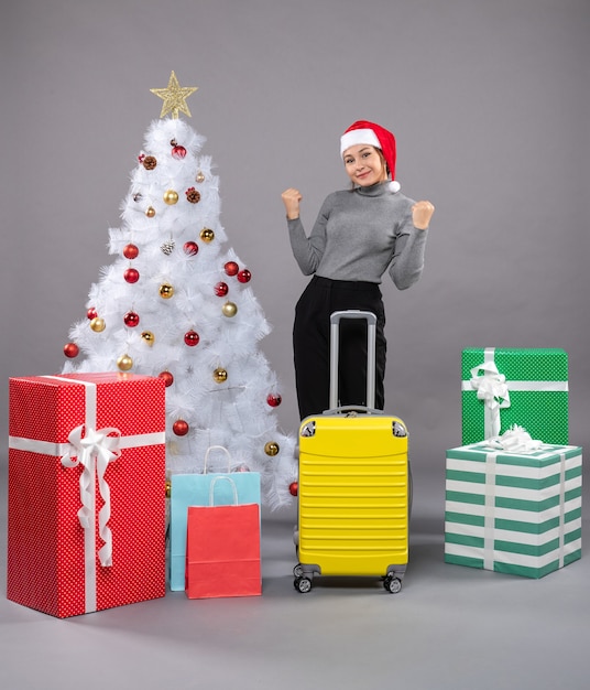 Woman wearing Santa Claus hat with luggage next to Christmas tree