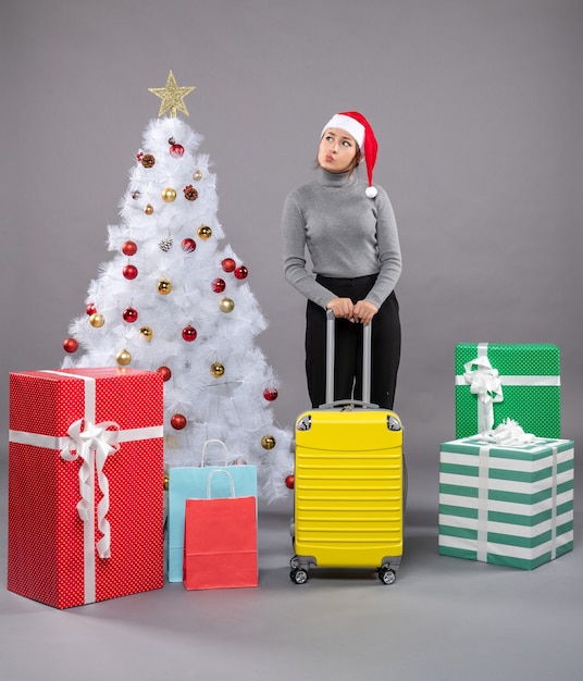 Woman wearing Santa Claus hat with luggage next to Christmas tree