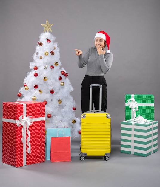 Woman wearing Santa Claus hat with luggage next to Christmas tree