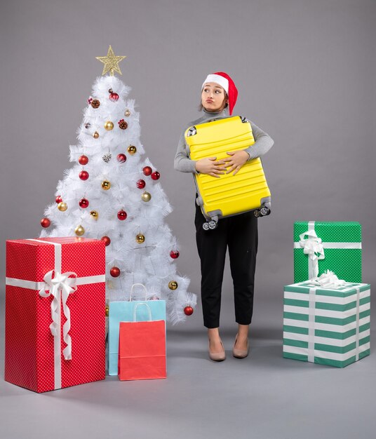 Woman wearing Santa Claus hat with luggage next to Christmas tree