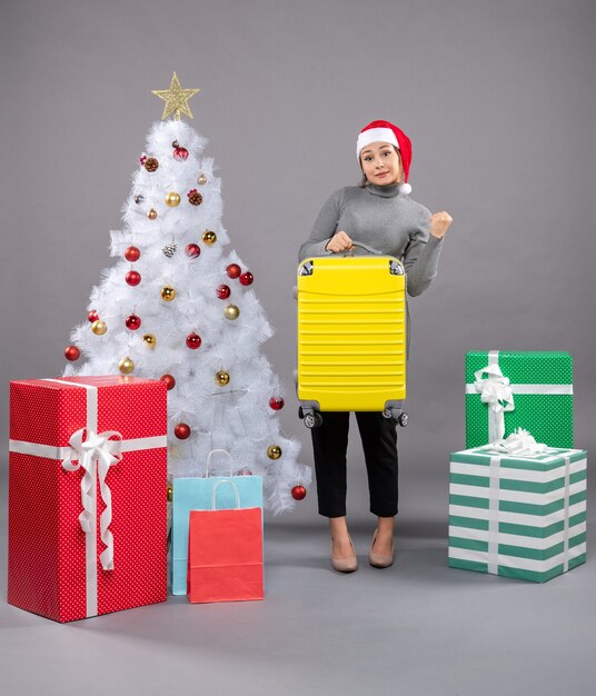 Woman wearing Santa Claus hat with luggage next to Christmas tree