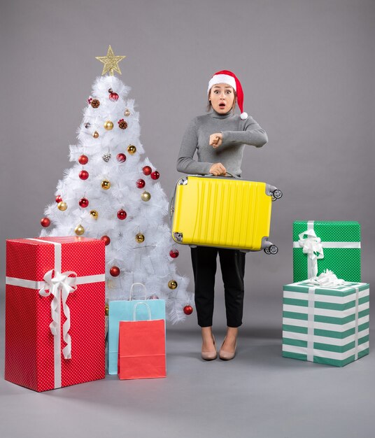 Woman wearing Santa Claus hat with luggage next to Christmas tree