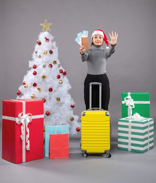 Woman wearing Santa Claus hat with luggage next to Christmas tree