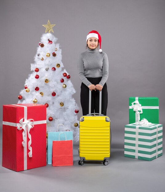 Woman wearing Santa Claus hat with luggage next to Christmas tree