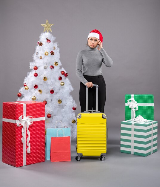 Woman wearing Santa Claus hat with luggage next to Christmas tree