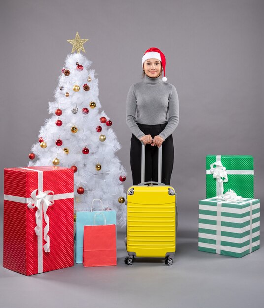 Woman wearing Santa Claus hat with luggage next to Christmas tree